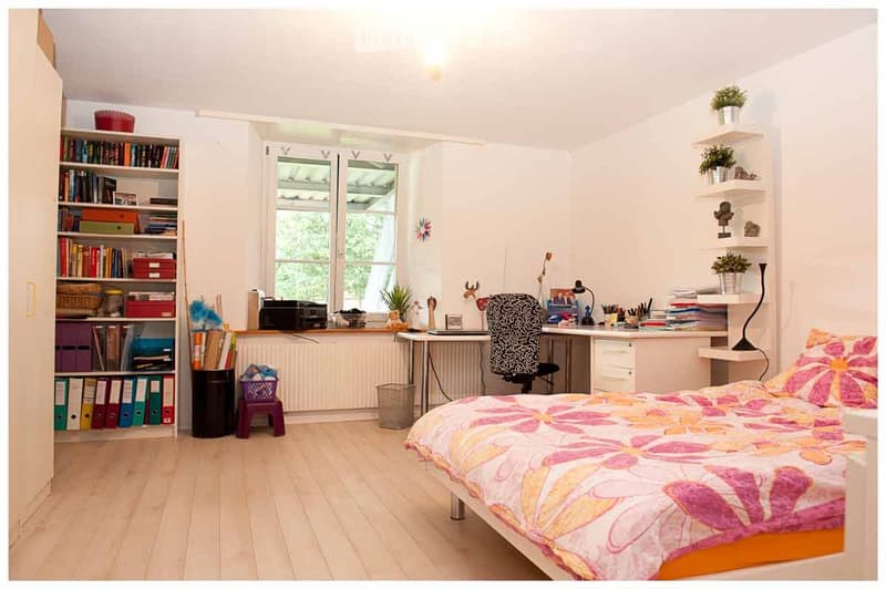 Bright bedroom with a bed, desk, bookshelf, and various decorative items. The room features wooden flooring, white walls, and large windows letting in natural light.