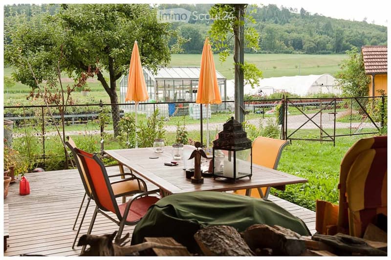 Wooden balcony with a table, chairs, and a lantern, overlooking a green field with greenhouses and trees.