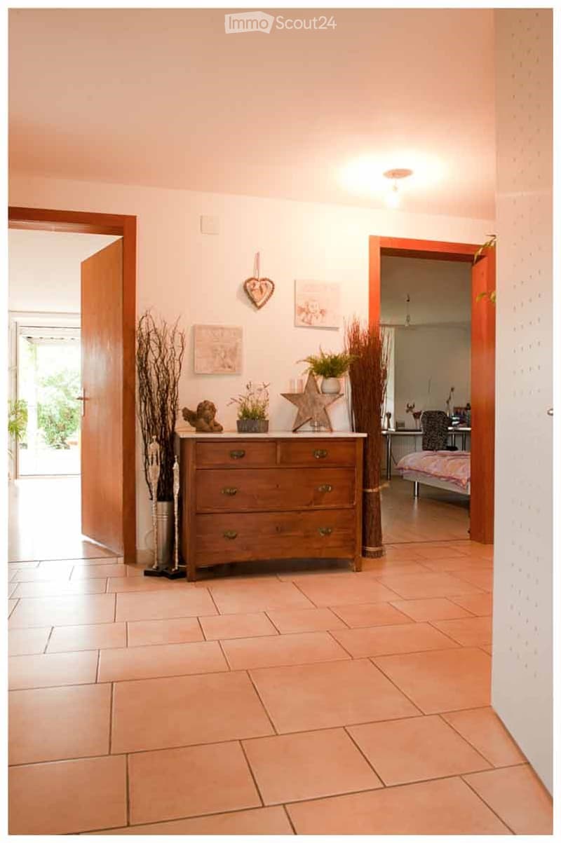 A well-lit hallway with tiled flooring, a wooden chest of drawers, decorative items including a star-shaped ornament and dried plants, and two open doorways leading to other rooms.