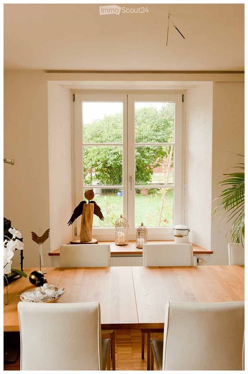 Bright dining room with large windows, wooden dining table, white chairs, decorative birdcages, and a wooden bird sculpture on a side table.