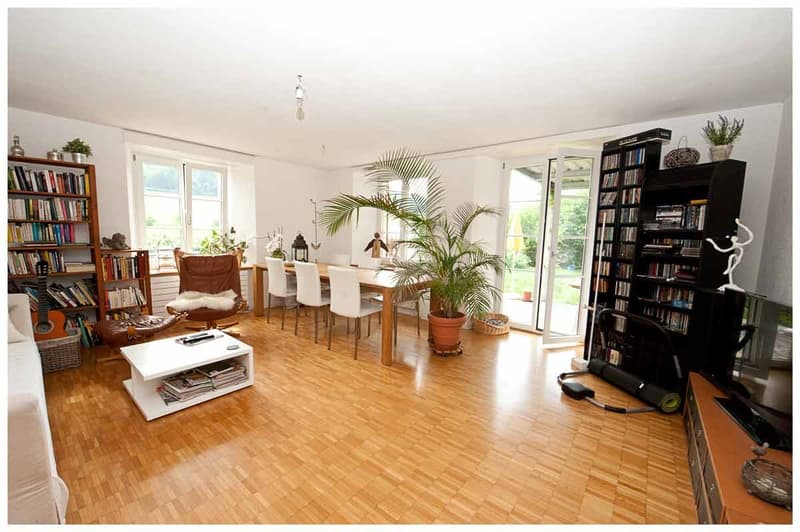 Bright, open living and dining area with wooden flooring, large windows, and modern furniture including a sofa, coffee table, dining table, chairs, bookshelves, and a guitar.