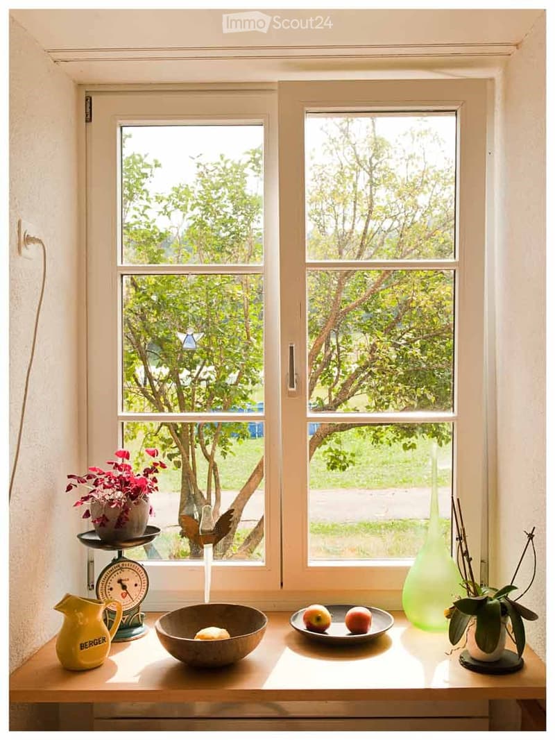 A small wooden table sits beneath a large window with a view of greenery. On the table are a green vase, a bowl with a fruit, a plate with apples, a yellow pitcher, and a scale with a flower pot on top.
