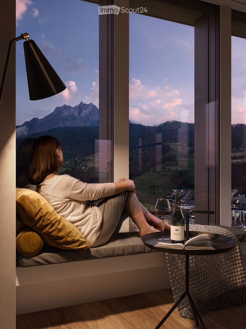 A person is sitting on a window seat with a view of mountains, holding a wine glass, with a bottle, book, and floor lamp nearby.