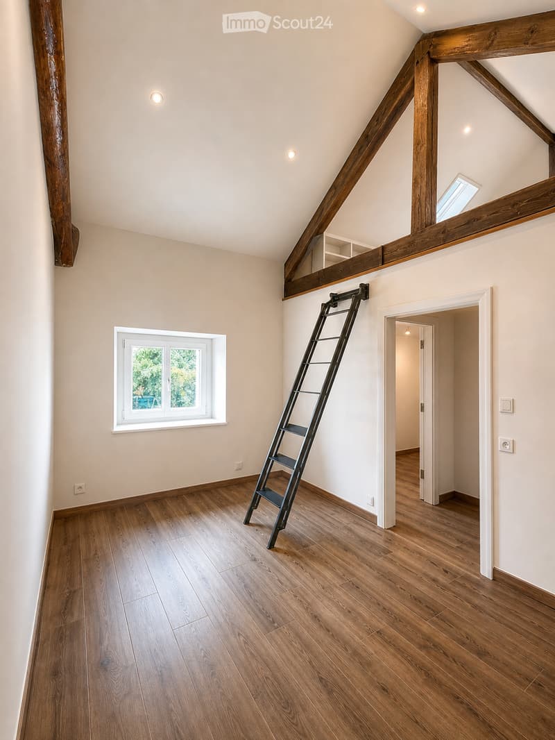 An empty attic room with wooden floors, exposed wooden beams, and a small window. A ladder is positioned against the wall, leading to a loft area.