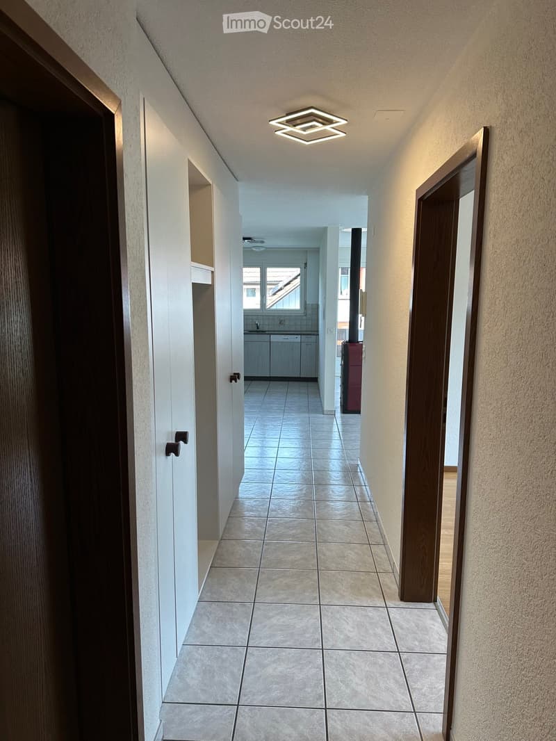 Clean hallway with tiled floor, white walls, and modern square ceiling light. Doors lead to adjacent rooms, including a kitchen area visible at the end.