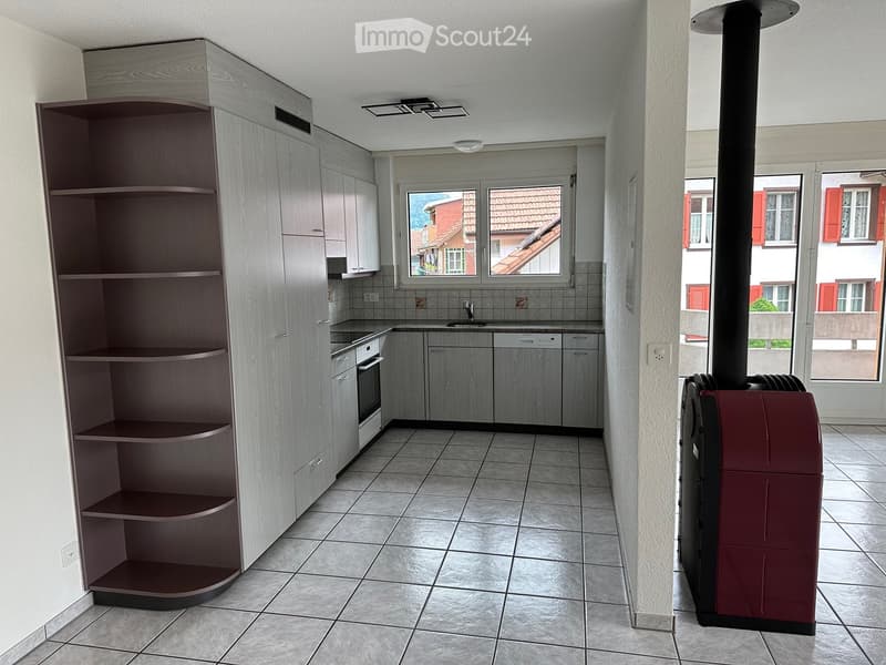 Modern kitchen with light wood cabinets, stainless steel appliances, tiled floor, and a large window overlooking a street with red-roofed buildings.