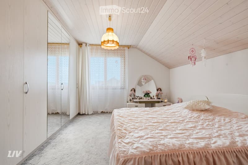 Cozy bedroom with a large bed, mirrored wardrobe, dresser, and decorative items on a small table. Natural light filters through sheer curtains.