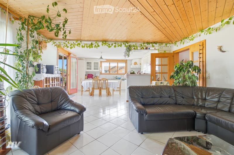Open-plan living and dining area with a black leather sofa, coffee table, dining table with chairs, and a kitchen area with cabinets and a counter.