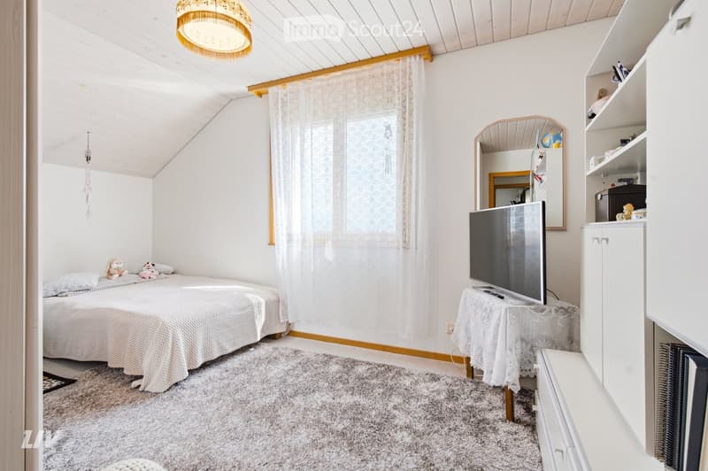 Cozy bedroom with a single bed, a small table with a TV, a mirror, and a cabinet. The room features a slanted wooden ceiling, a window with white curtains, and a carpeted floor.