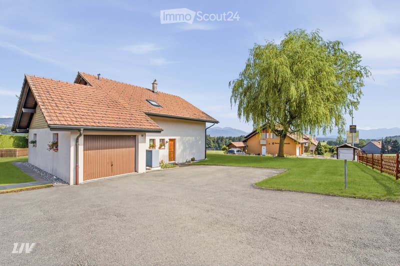 Two-story house with a red-tiled roof, beige walls, and a wooden garage door. The house features a small porch with a wooden door and flower boxes, surrounded by a well-maintained lawn and a large weeping willow tree.