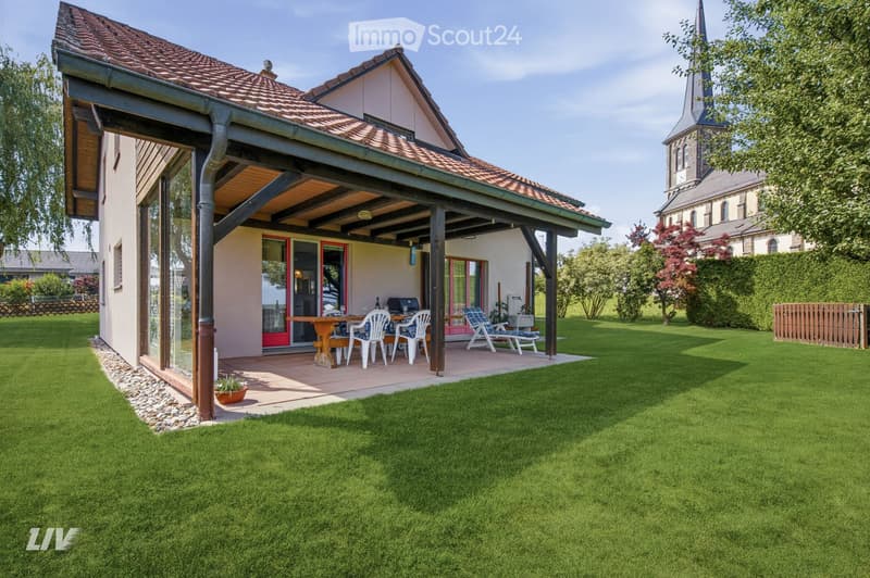 A well-maintained garden with a covered patio area, outdoor furniture, and a barbecue. The house has a modern design with large windows and a tiled roof.