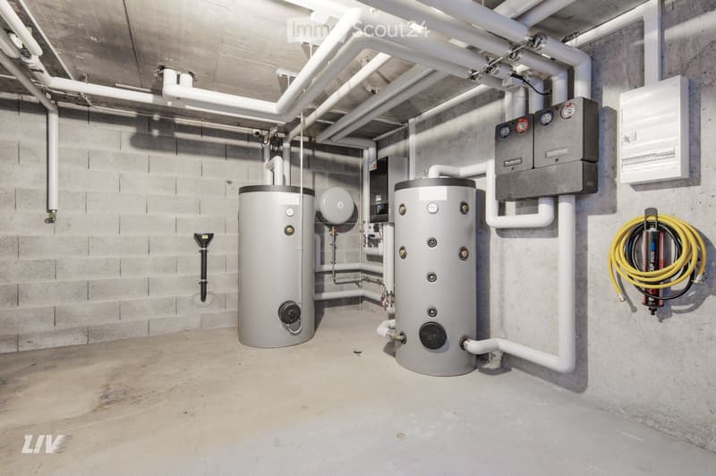 Utility room with two water heaters, control panel, exposed pipes, and coiled cable on wall.
