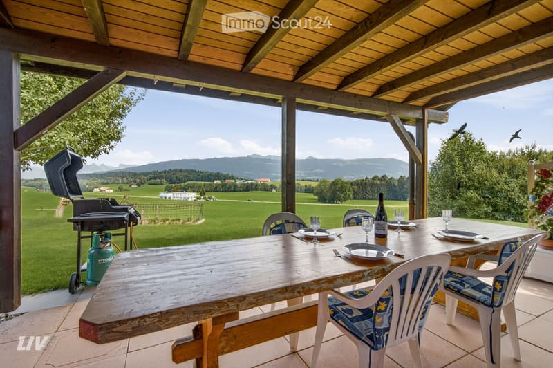A wooden outdoor dining area with a table, chairs, and a grill, overlooking a scenic valley with mountains in the background.