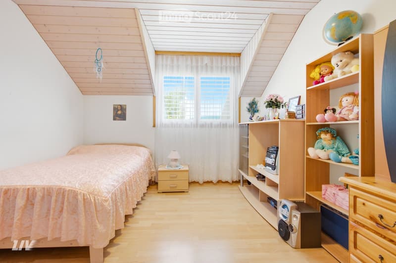 Cozy bedroom with slanted wooden ceiling, bed with pink bedspread, nightstand with lamp, wooden shelves with toys and a globe, and a window with white curtains.