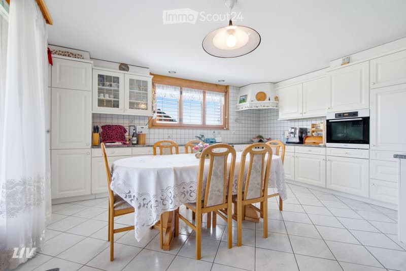 Bright kitchen with white cabinets, tiled floor, and a dining area featuring a round table with six chairs. The kitchen includes an oven, a refrigerator, and various small appliances on the counter.