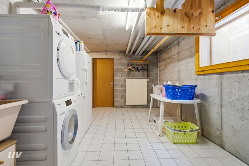Laundry room with white tiled floor, washing machine, dryer, small table with baskets, radiator, and a door leading to another area.