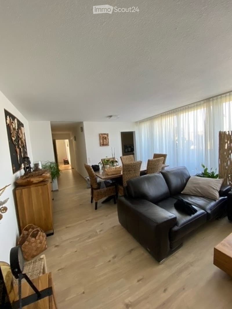 Bright living and dining area with hardwood flooring, large windows with white blinds, black leather sofa, dining table with wicker chairs, sideboard, and decorative plants.