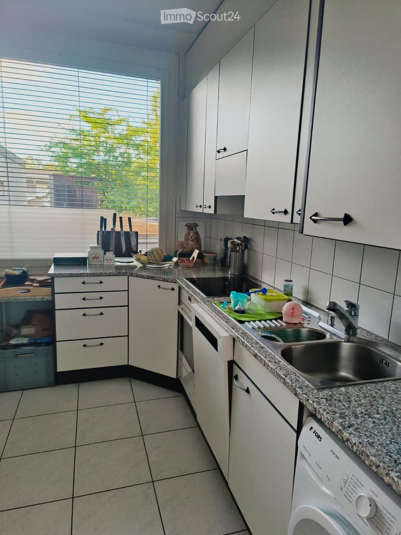 Modern kitchen with white cabinets, granite countertop, stainless steel sink, and various appliances including a dishwasher and oven. The space is well-lit with large windows and features a variety of kitchen items on the counter.