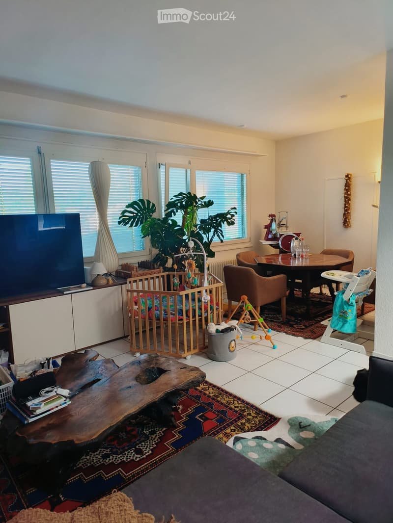 A well-lit living room with a large window, a wooden coffee table, a sofa, a baby crib, a dining table with chairs, and a plant in the corner.