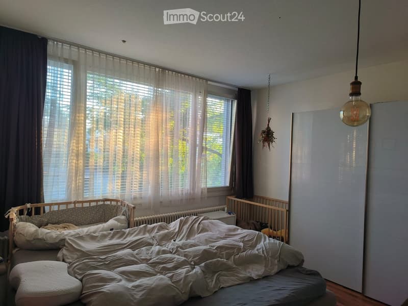 A cozy bedroom with a large window, a bed with rumpled bedding, a small wooden cradle, and a hanging decorative plant.