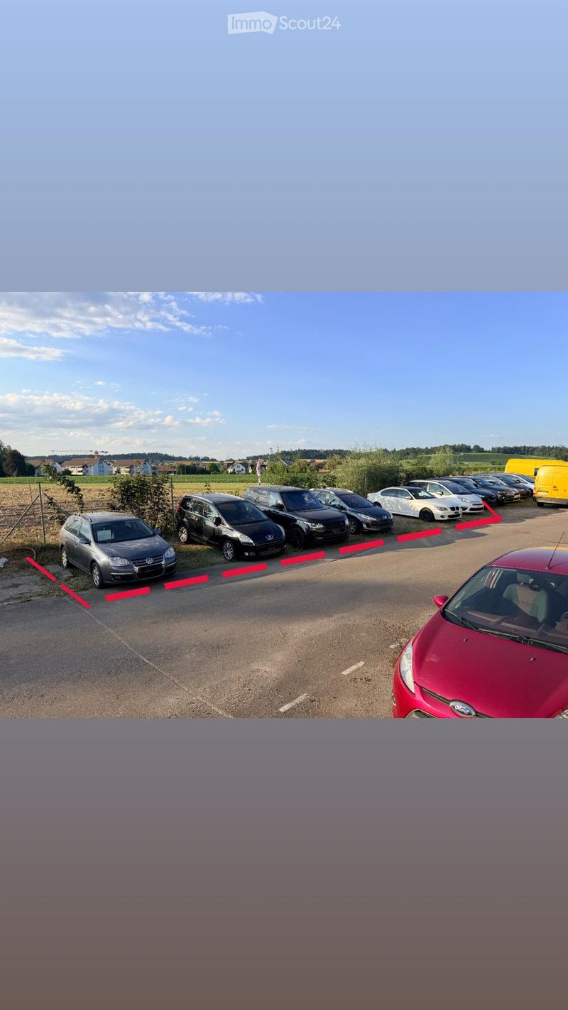 A parking lot with several cars parked in rows, surrounded by a rural landscape with fields and houses in the background.