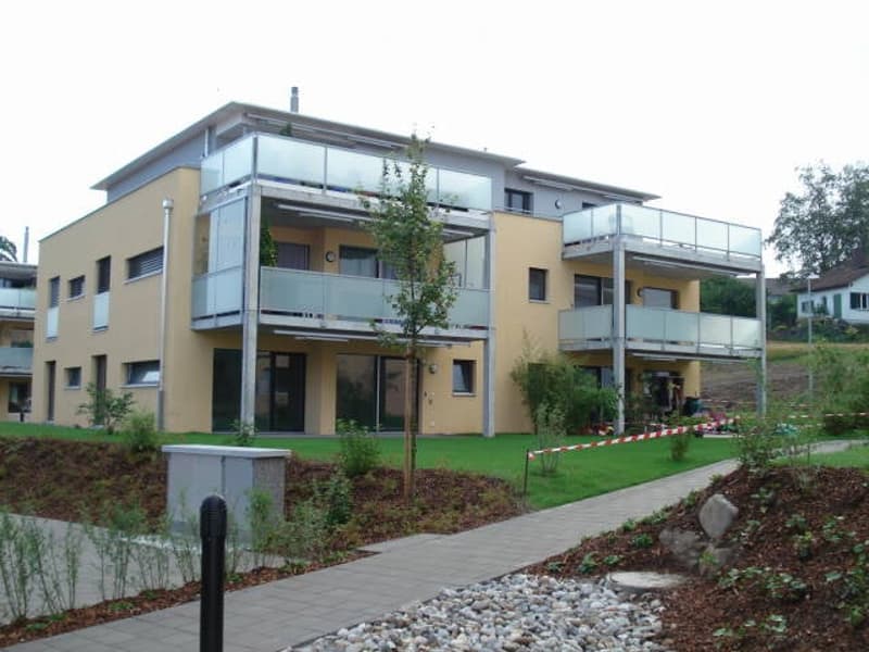 Modern two-story building with yellow and white facade, large glass balconies, and landscaped surroundings.