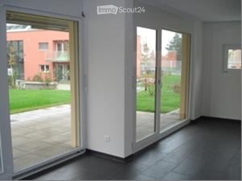 Empty room with large sliding glass doors leading to a patio, dark tiled floor, white walls, and a view of a green lawn and neighboring building.