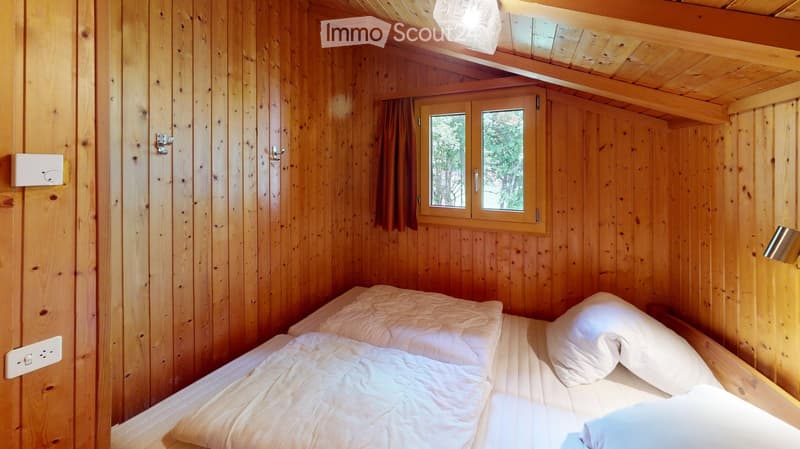 Cozy wooden bedroom with a double bed, white bedding, and a window with curtains. The room features exposed wooden beams and a warm, natural aesthetic.