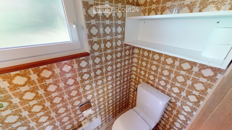 Bathroom with vintage patterned tiles, a white toilet, and a built-in shelf above it. The room features a window with white frames and a wooden trim along the lower wall.