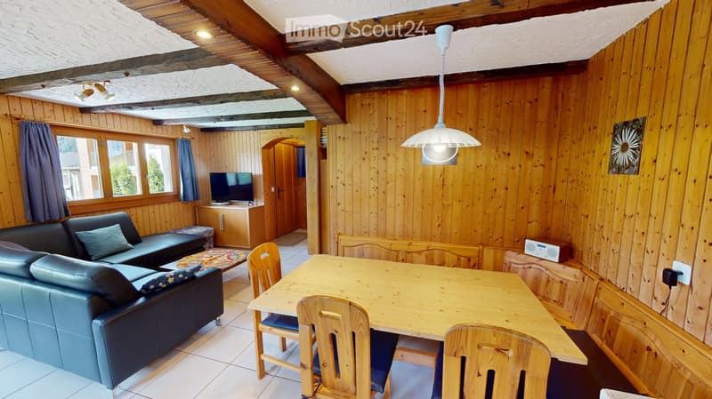 A cozy living room with wooden walls and exposed wooden beams, featuring a dark blue leather sofa, a wooden dining table with chairs, a television, and a hanging lamp.