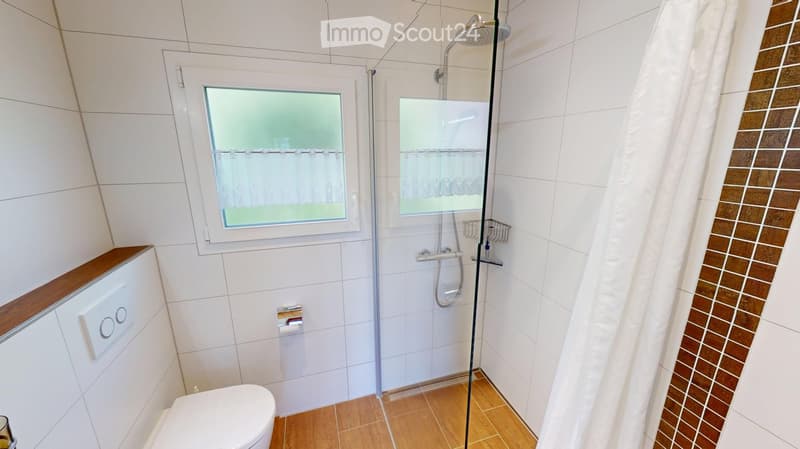 Modern bathroom with white tiled walls, wooden floor, toilet, shower with glass enclosure, and a window with frosted glass.