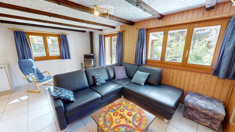 Cozy living room with wooden panel walls, exposed wooden beams, large windows with blue curtains, a dark blue sectional sofa with decorative pillows, a colorful coffee table, a wooden chair, and an ottoman.