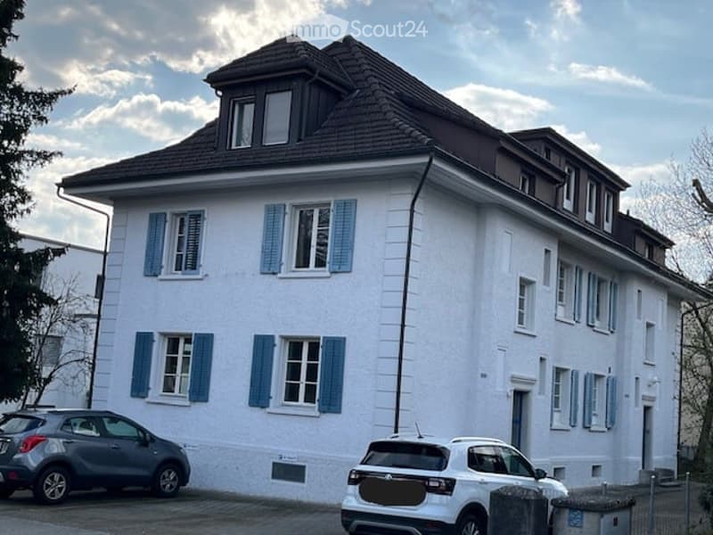 A two-story white building with blue shutters and a dark roof, featuring multiple windows and a small dormer window on the roof.