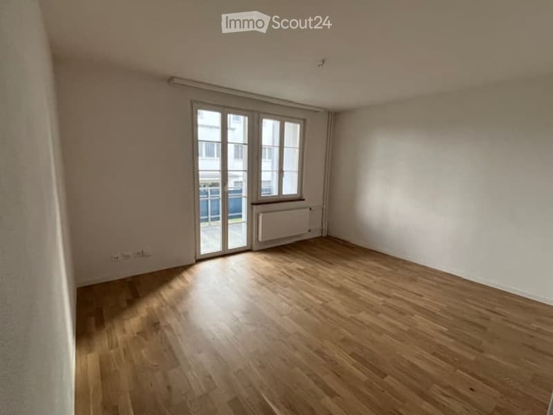 Empty room with wooden flooring, large window with sliding glass door, radiator, and visible electrical outlets.