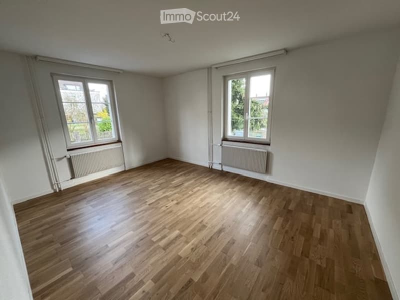Empty room with wooden flooring, white walls, two windows, and radiators.