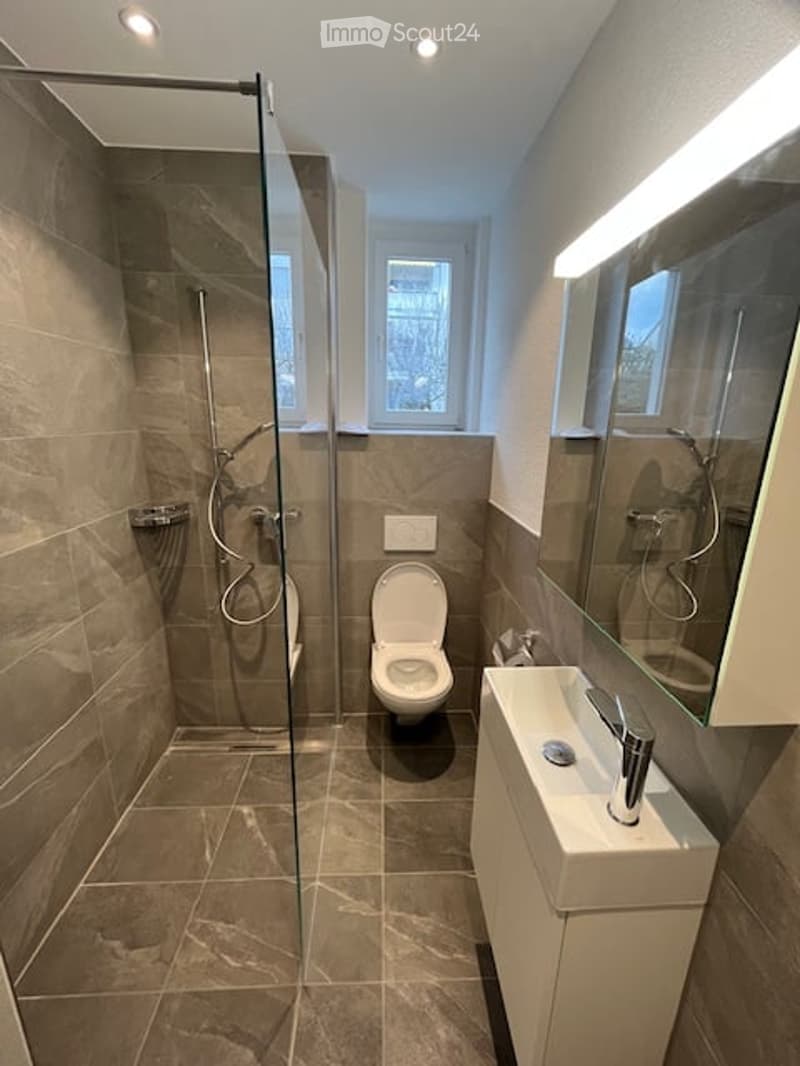 Modern bathroom with a glass-enclosed shower, toilet, sink, and large mirror. The walls and floor are tiled in a neutral brown color, and the space is well-lit with recessed lighting.