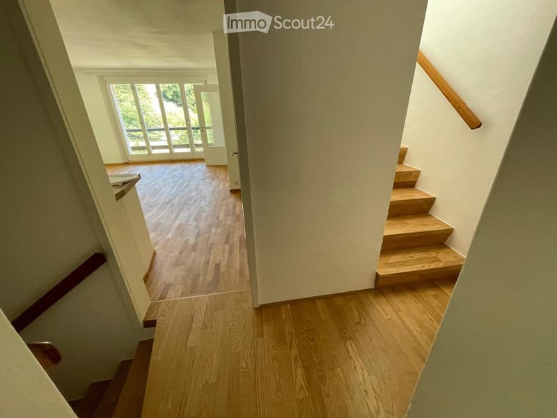 A well-lit hallway with wooden flooring and a staircase leading upwards, featuring a wooden handrail and a large window at the end.