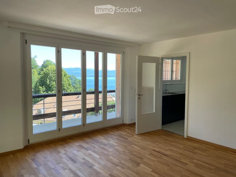 Empty room with wooden flooring, large sliding glass doors leading to a balcony, and an open doorway to a small kitchen area.