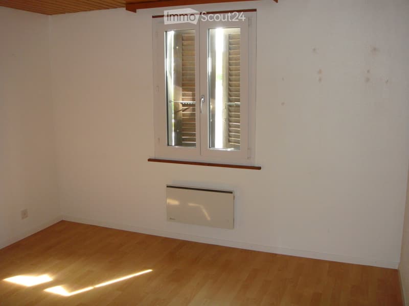 Empty room with light wooden flooring, white walls, a double window with blinds, a radiator, and a small shelf above the window.