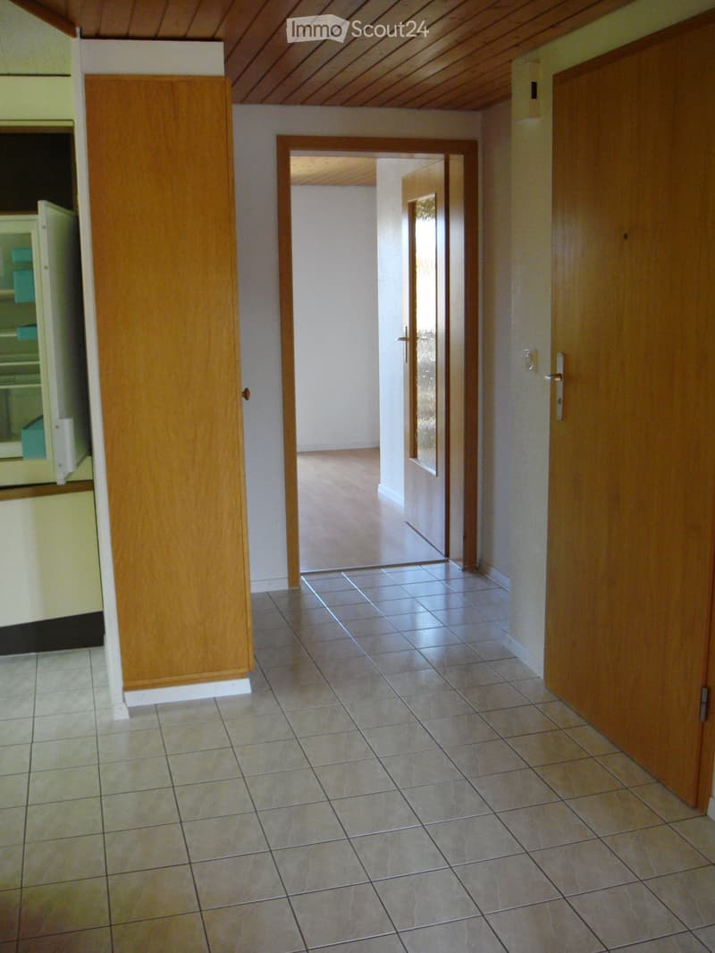 Empty hallway with tiled floor, wooden doors, and a visible kitchen area on the left side.