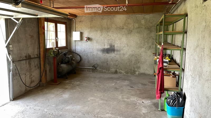 Empty garage with concrete floor, exposed wooden beams, and a single window. Contains a metal shelving unit, air compressor, and a few storage items.