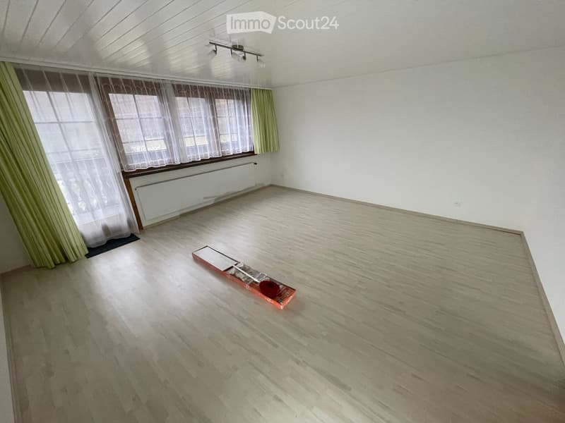 Empty room with wooden flooring, white walls, large window with curtains, radiator, and a small box on the floor.