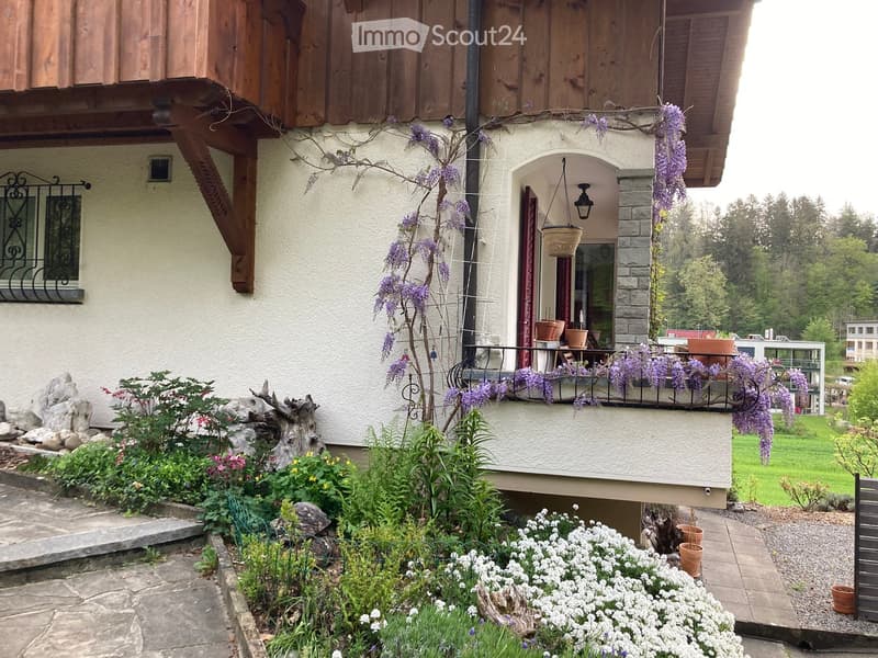 A well-maintained garden with a variety of flowers, shrubs, and a small tree with purple blossoms. The garden features a stone pathway, decorative rocks, and a wooden deck with potted plants and hanging baskets.