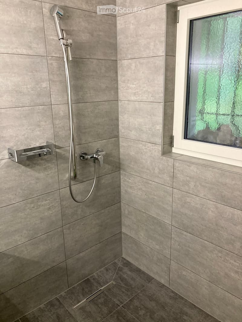 Modern shower area with grey tiled walls and floor, chrome showerhead, faucet, and handrail.