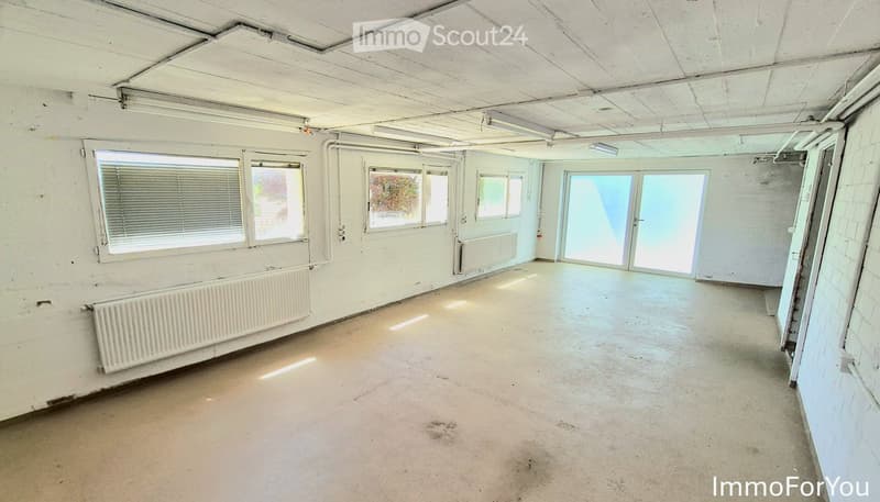 Empty room with bare walls, concrete floor, and large windows with blinds. Radiators are visible along the walls.