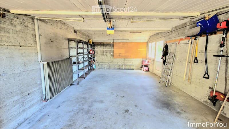 Empty garage with concrete floors and walls, metal shelving, and various tools and equipment stored on the walls and floor.