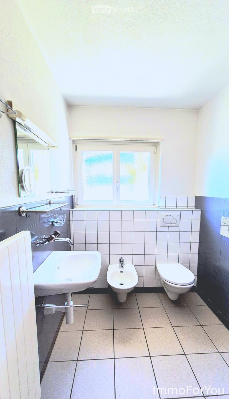 Modern bathroom with a sink, toilet, bidet, and mirror. Light-colored tiles and a window provide natural light.