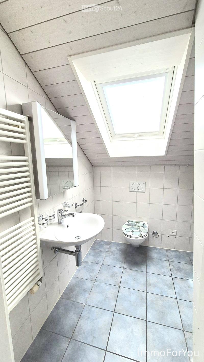 Modern bathroom with a white sink, chrome faucet, toilet with a decorative stone seat cover, a white radiator, and a large mirror. The room features grey tiled walls and floor, and a skylight window.