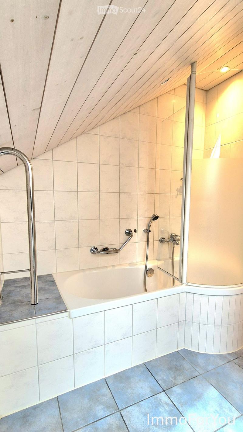 Modern bathroom with a bathtub, handheld showerhead, and handrail, featuring light-colored tiles and a sloped ceiling.