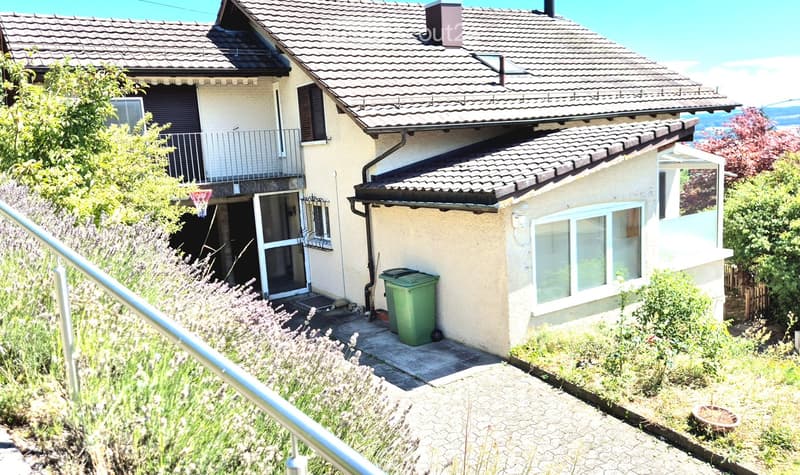 Two-story house with a tiled roof, beige exterior, and a small balcony on the upper level. The house has multiple windows, a green trash bin, and is surrounded by greenery and a paved pathway.